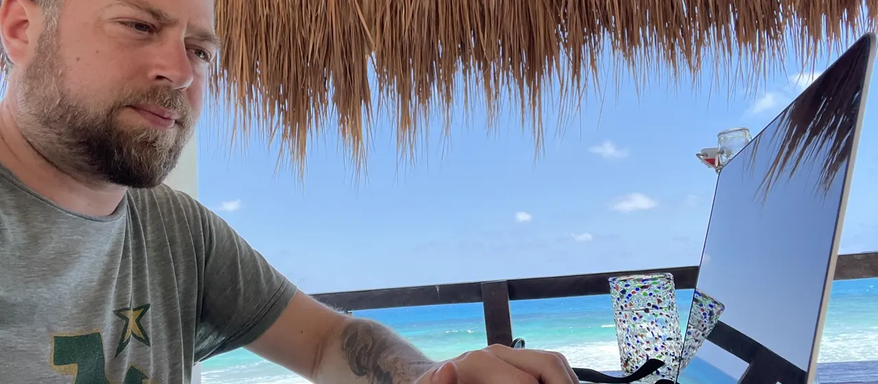 Jared working on a MacBook under a thatched palapa, ocean and beach in the background.