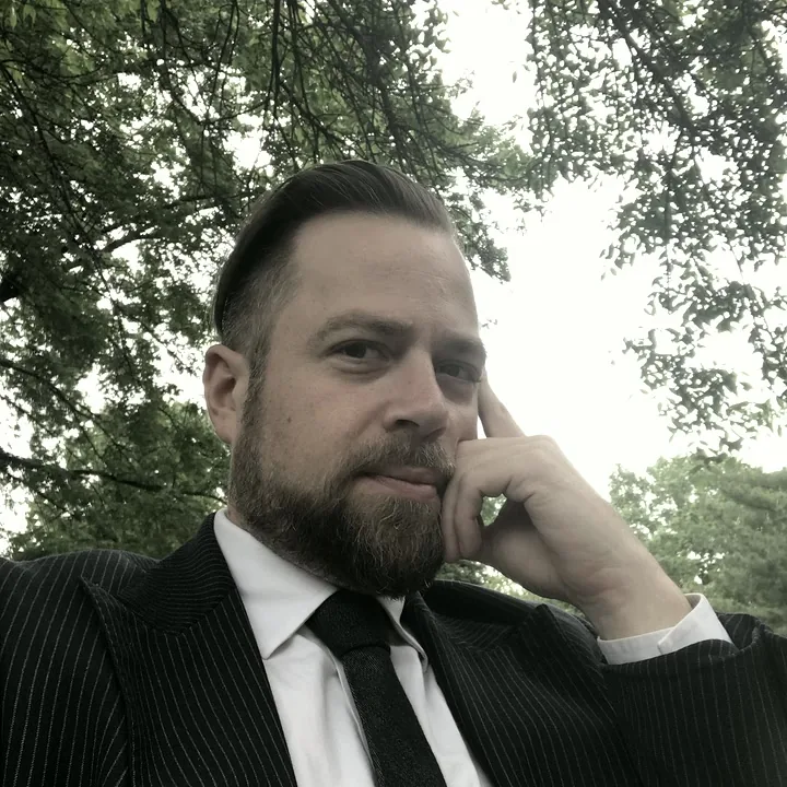 Jared in a pinstripe suit, contemplative, tree canopy overhead.